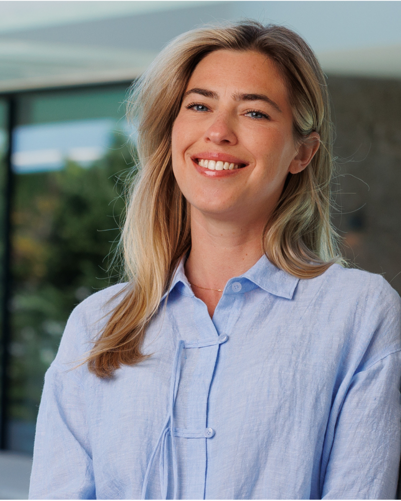 Smiling blond woman wearing a light blue blouse, standing outdoors near a modern building.