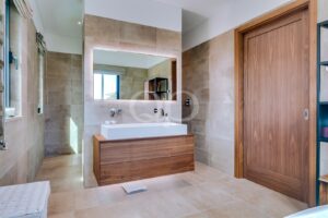 Salle de bains moderne avec un meuble-lavabo en bois, un lavabo rectangulaire blanc, un grand miroir, des murs et un sol carrelés de couleur beige, ainsi qu'une porte en bois sur la droite.