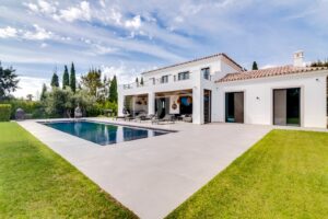 Une villa blanche à deux étages avec un toit en tuiles de terre cuite, un coin salon au bord de la piscine et une pelouse impeccablement entretenue sous un ciel bleu.