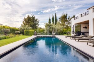Vue depuis la piscine d'une maison moderne blanche, avec des chaises longues disposées le long d'une piscine rectangulaire bleue et un jardin paysager en arrière-plan.
