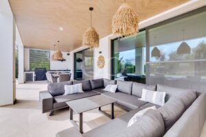 Une terrasse extérieure couverte avec un canapé modulable gris, des coussins rayés et des suspensions en osier suspendues sous un plafond en bois.