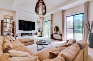 Un salon spacieux et moderne, avec un canapé d'angle beige, une télévision fixée au mur et des baies vitrées donnant sur un jardin ensoleillé.