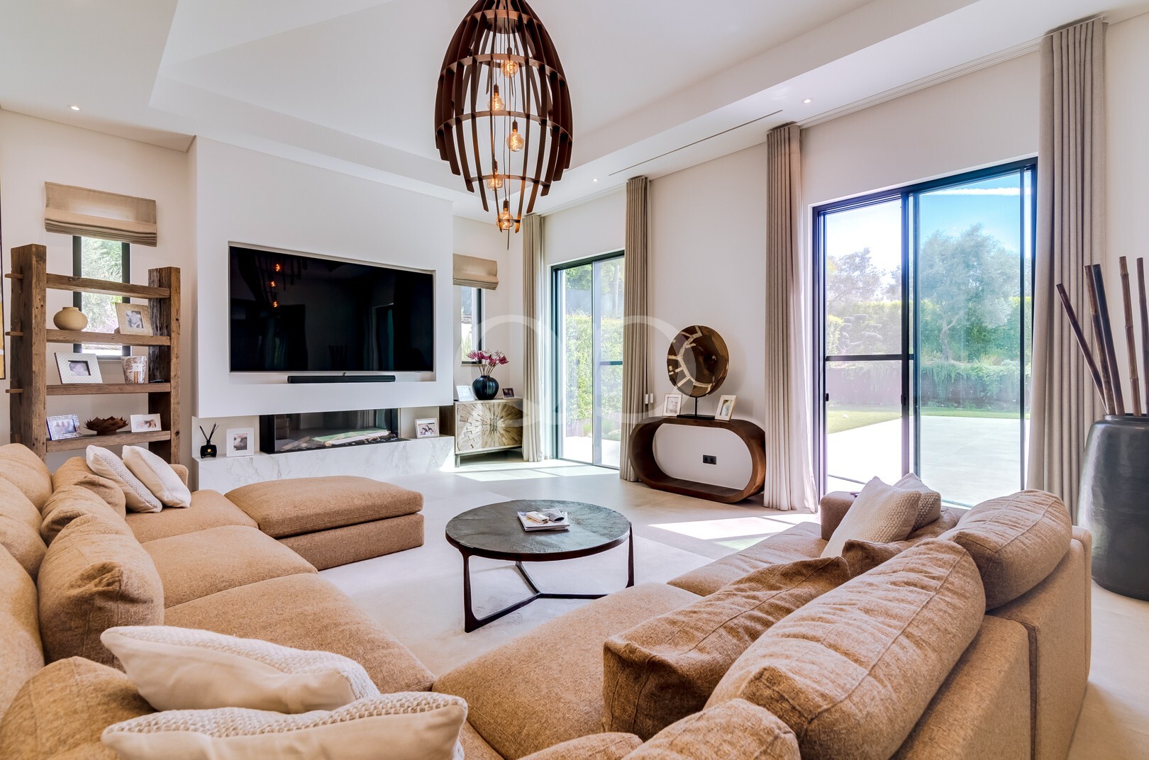 Un salon spacieux et moderne, avec un canapé d'angle beige, une télévision fixée au mur et des baies vitrées donnant sur un jardin ensoleillé.