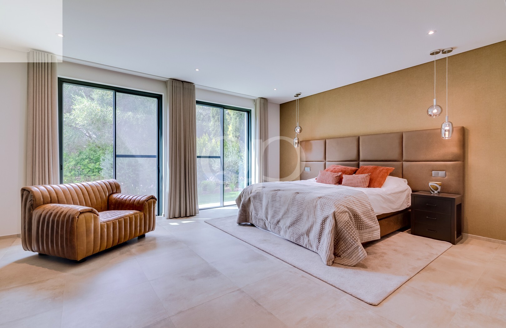 Chambre moderne avec un lit king size, une tête de lit capitonnée beige, des suspensions et un canapé en cuir marron près de grandes baies vitrées.