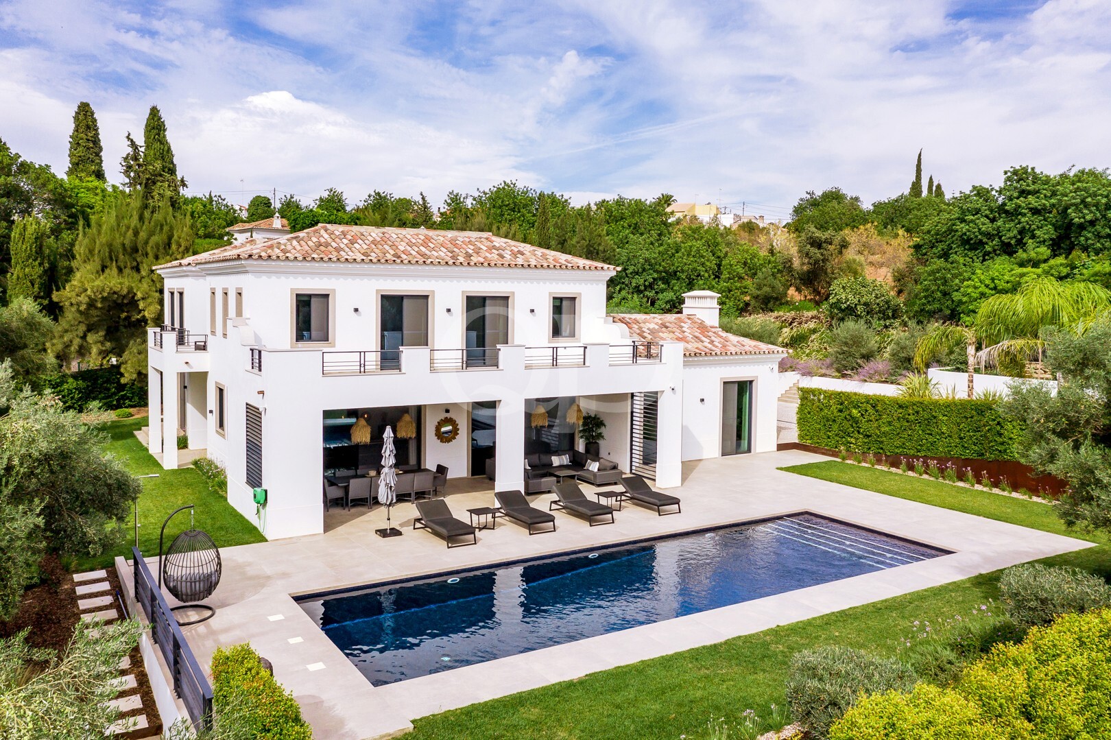 White two-story villa with terracotta roof, a spacious patio and a rectangular pool surrounded by loungers and a seating area.