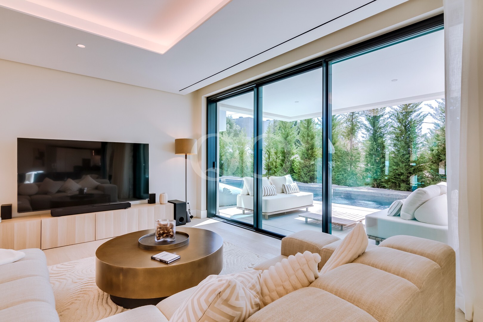 Bright, modern living room with a wall-mounted TV, cream sofa, and a round coffee table, opening to a patio via large glass sliding doors and pool seen outside.