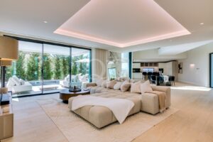 Bright, open-plan living room with a large beige sectional, round coffee table, and a cream rug, opening to a poolside garden through sliding glass doors, creating outdoor views.