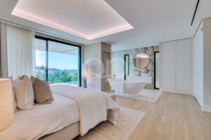 Bright, modern bedroom with a freestanding white tub in an open-plan bathroom and a large glass door showing a hillside view.