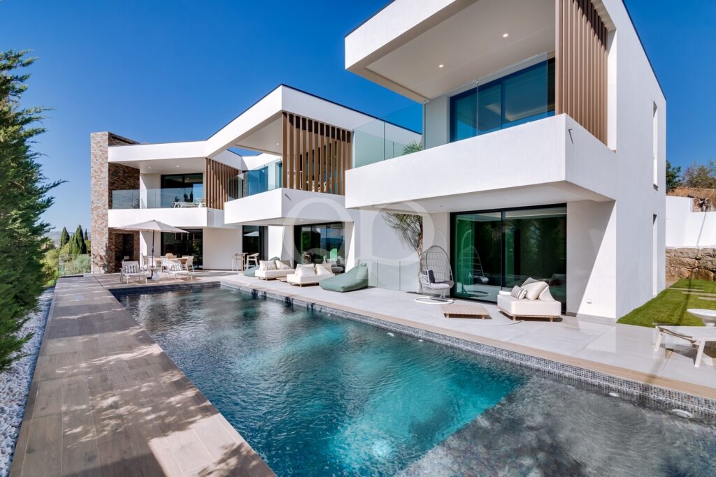 Contemporary white villa with glass balconies, wooden slat screens, and a long rectangular pool by an outdoor lounge area.