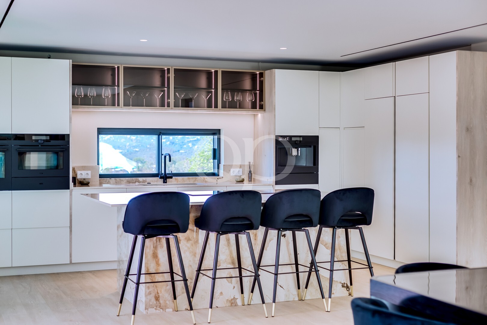Modern white kitchen with marble island, four black bar stools, and built-in ovens; glass-front cabinets above a window showing mountains.