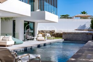 Modern white villa with a rectangular pool, beige loungers, and a hanging egg chair on a sunlit patio under a blue sky.