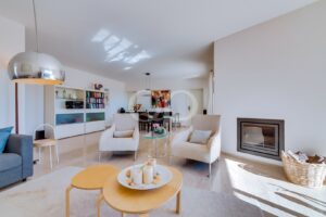 Bright open-plan living room with beige armchairs, glass coffee table, and a right-side fireplace; dining area in the back.