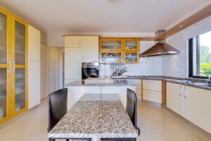 Bright modern kitchen with granite island, cream cabinets, and a stainless range hood by a large window.