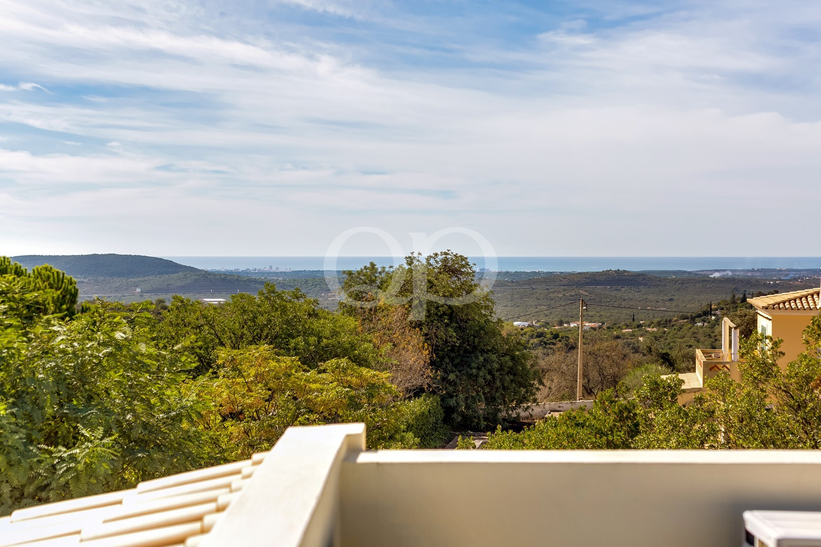 Broad coastal landscape with rolling hills, greenery in foreground, and distant sea under a blue sky with wispy clouds (view from a terrace).