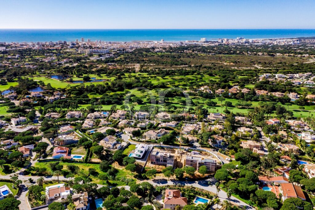 Vista aérea de uma cidade costeira com um grande campo de golfe verdejante e bairros residenciais, com o mar no horizonte.