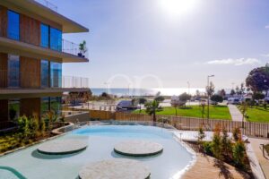 Modernes Mehrfamilienhaus mit Glasbalkonen und Blick auf einen Pool sowie eine sonnige Strandlandschaft jenseits des Meeres.
