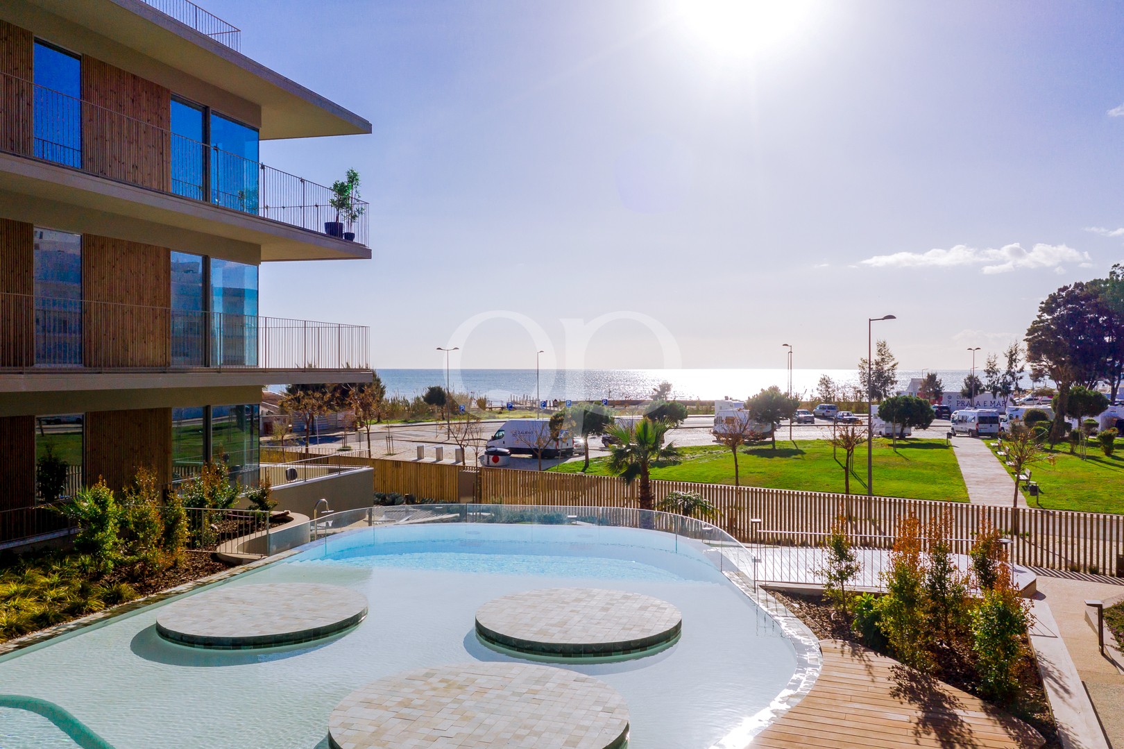 Modernes Mehrfamilienhaus mit Glasbalkonen und Blick auf einen Pool sowie eine sonnige Strandlandschaft jenseits des Meeres.