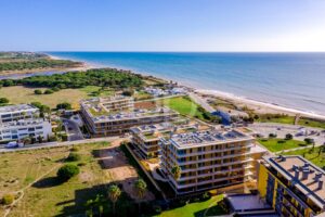 Luftaufnahme eines Strandkomplexes mit modernen weißen Gebäuden, grünen Parks und einem Sandstrand, der von oben betrachtet auf das ruhige blaue Meer trifft.