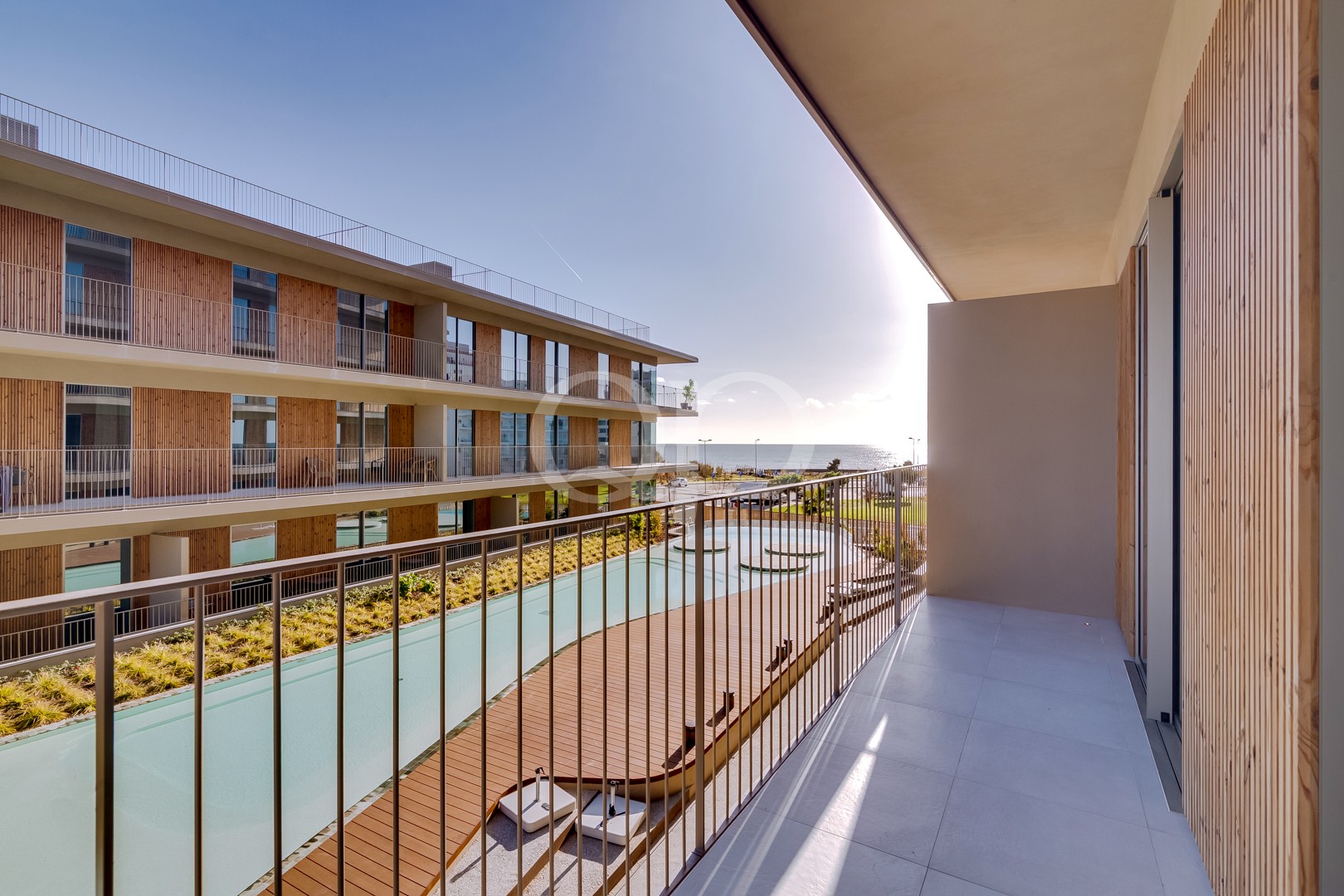 Blick vom Balkon auf den Innenhof eines Ferienresorts mit Pool, Holzgeländern und dem Meer in der Ferne.