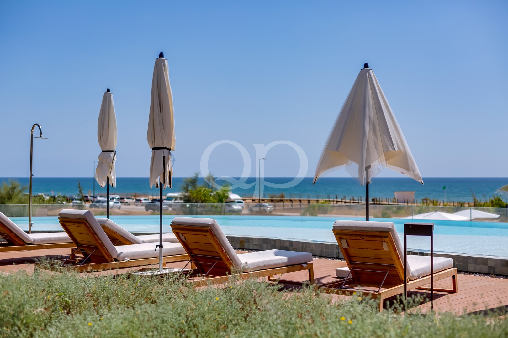 Liegestühle am Pool mit eingeklappten Sonnenschirmen, mit Blick auf das ruhige Meer unter einem strahlend blauen Himmel am Poolbereich eines Resorts.