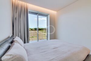 Schlafzimmer mit einer großen Glasschiebetür, die auf einen Balkon führt; zartgraue Vorhänge rahmen den Blick auf die grasbewachsene Landschaft und die Bäume draußen ein.