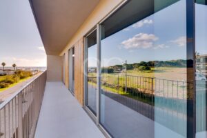Moderner Balkon an einem Gebäude mit Glasfassade, weißer Fliesenboden und Metallgeländer, mit Blick auf eine Küstenlandschaft mit Palmen und strahlend blauem Himmel.