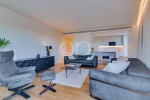 Ein offen gestaltetes Wohnzimmer mit grauen Sofas, einem passenden Sessel und einer Ottomane sowie einem weißen Zottelteppich, das in eine minimalistisch gestaltete Küche mit einer Frühstückstheke übergeht.
