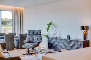 Ein modernes Wohnzimmer mit einem grauen Drehsessel, passenden Liegesesseln und einem niedrigen Couchtisch auf einem hellen Teppich, neben einem großen Fenster mit weißen Vorhängen. Ein schwarzes Sideboard mit geometrischem Muster und eine hohe Pflanze runden die Einrichtung ab. Der Raum wird von natürlichem Licht durchflutet.