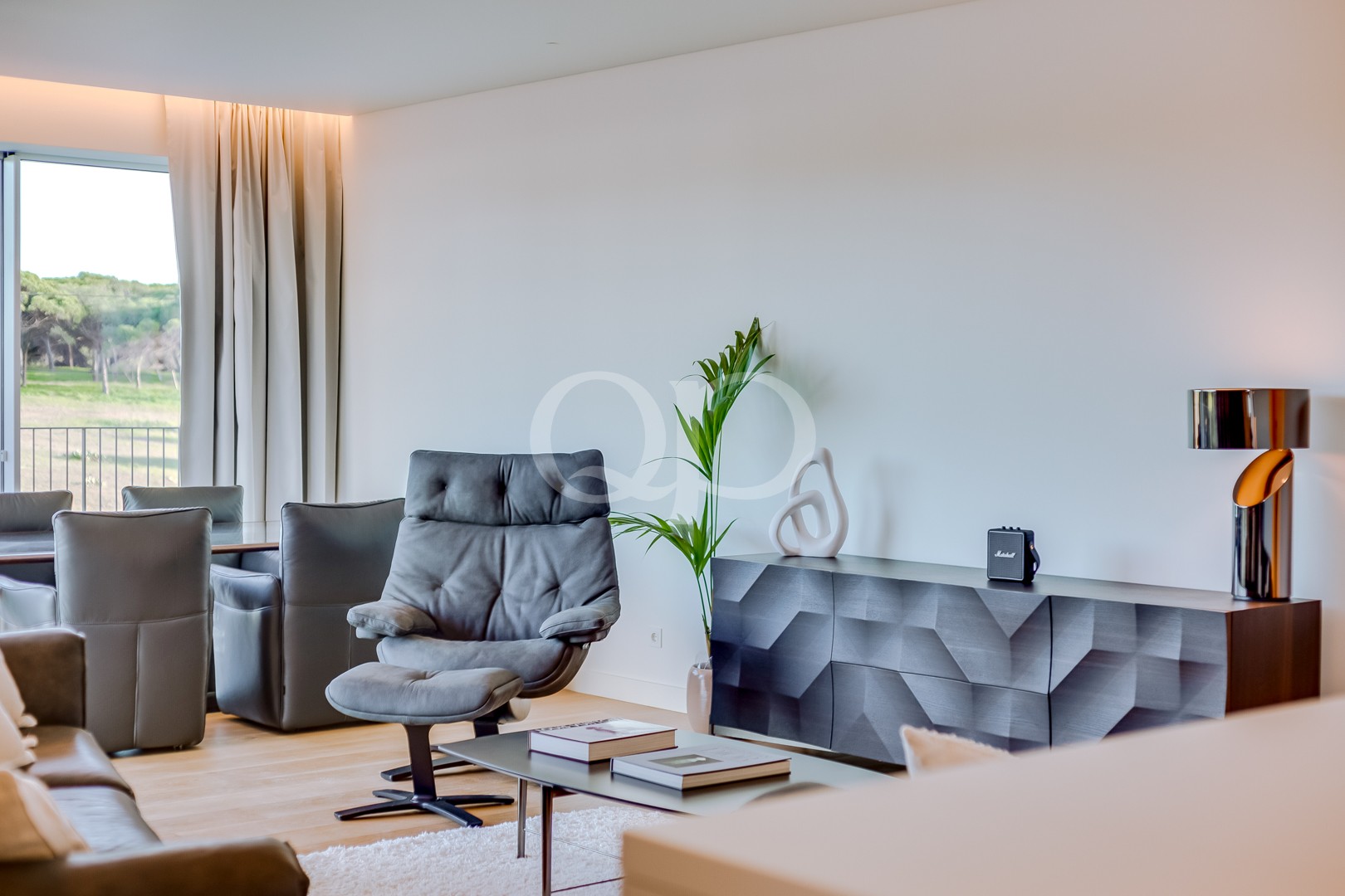 Ein modernes Wohnzimmer mit einem grauen Drehsessel, passenden Liegesesseln und einem niedrigen Couchtisch auf einem hellen Teppich, neben einem großen Fenster mit weißen Vorhängen. Ein schwarzes Sideboard mit geometrischem Muster und eine hohe Pflanze runden die Einrichtung ab. Der Raum wird von natürlichem Licht durchflutet.