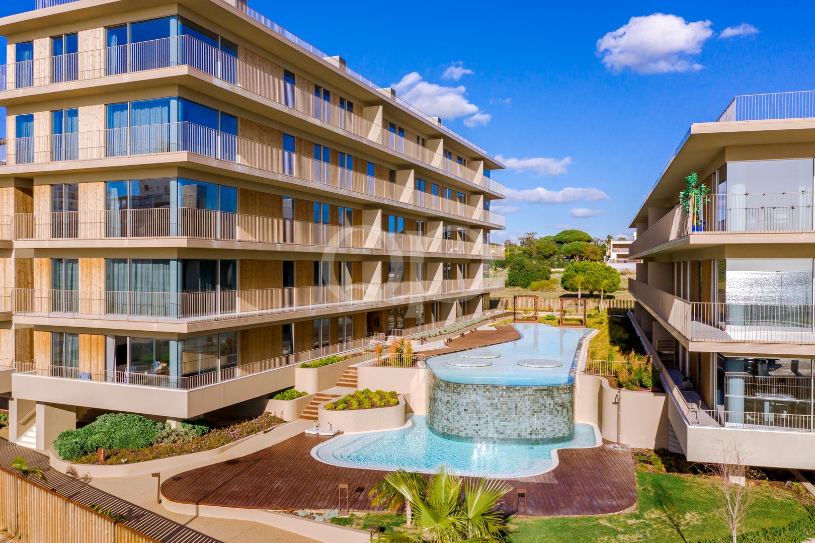 Modern apartment complex with multiple balconies surrounding a winding pool courtyard under a blue sky
