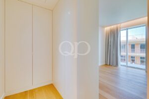 Bright hallway with white built-in wardrobes on the left, leading to a sunlit living area with large glass doors and gray curtains.