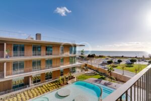 Modern seaside apartment building with glass balconies and a curved pool courtyard overlooking the ocean on a sunny day.