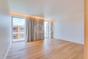 Bright empty living room with light wood floors, white walls, and large windows with gray curtains letting in natural light.