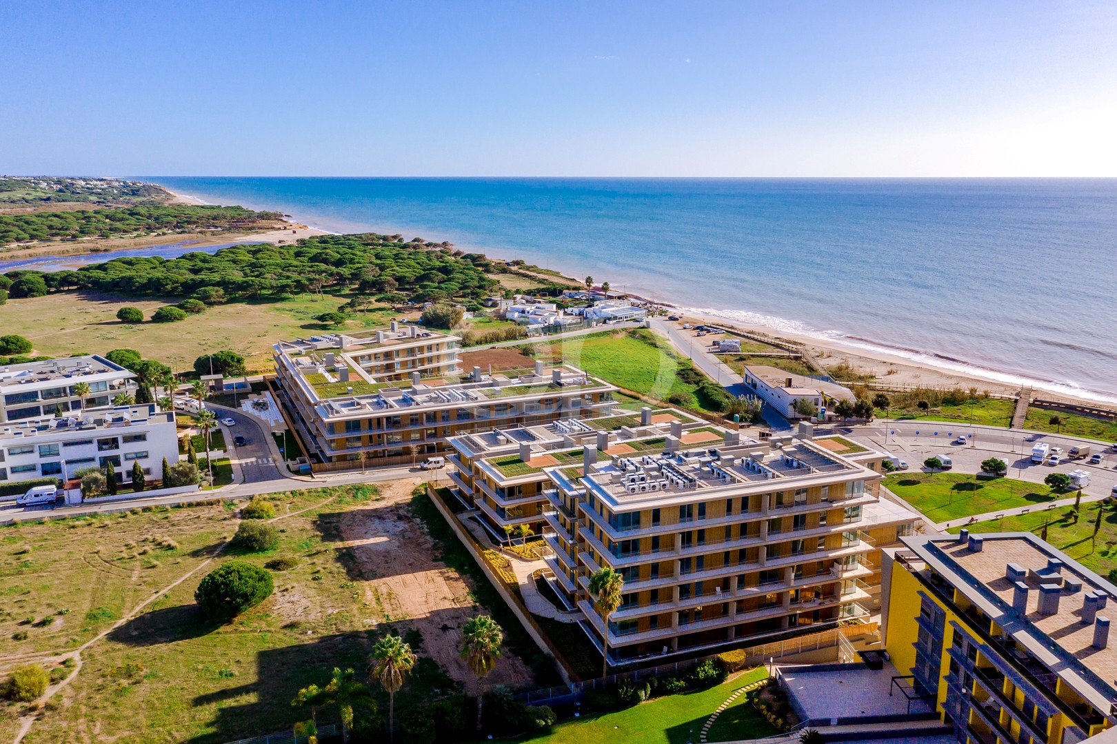 Aerial view of modern beachfront apartments facing a sandy beach and blue sea, with green lawn and trees nearby.