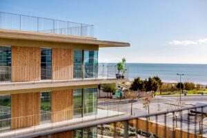 Modern multi-story building with wood-paneled balconies overlooking a coastal street and the sea under a clear blue sky, during daytime.