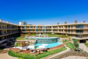 Pátio de um hotel moderno com uma piscina circular no centro, espreguiçadeiras e cabanas à sombra, sob um céu azul límpido.