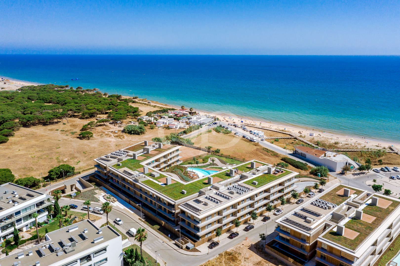 Aerial view of a coastal resort with a turquoise beach, blue sea, and a modern apartment complex with a rooftop pool and green gardens.