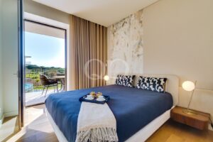 Quarto de hotel com colcha azul-marinho, manta bege e duas almofadas com padrões em azul-marinho; porta de vidro que dá para a varanda, com mesa e cadeiras ao ar livre e vegetação ao fundo.