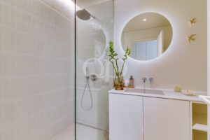 Casa de banho minimalista com uma cabina de duche com paredes de vidro, um espelho redondo retroiluminado e um lavatório branco com um vaso de bambu.
