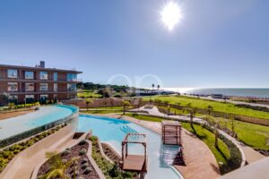 Resort com uma piscina exterior curva, um deck de madeira e vista para o mar, sob um sol radiante.