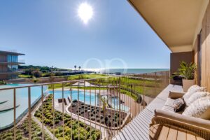 Varanda com assentos em bege e um vaso de plantas, com vista para a piscina de um resort e para o oceano num dia de sol