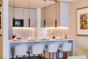 Cozinha moderna com armários brancos, revestimento em mármore e um balcão de pequeno-almoço com pratos e copos dispostos sob candeeiros suspensos.