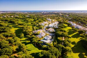 Vue aérienne d'un complexe de golf en bord de mer, avec des maisons modernes blanches nichées au milieu de fairways verdoyants et d'une végétation dense, l'océan se profilant à l'horizon.