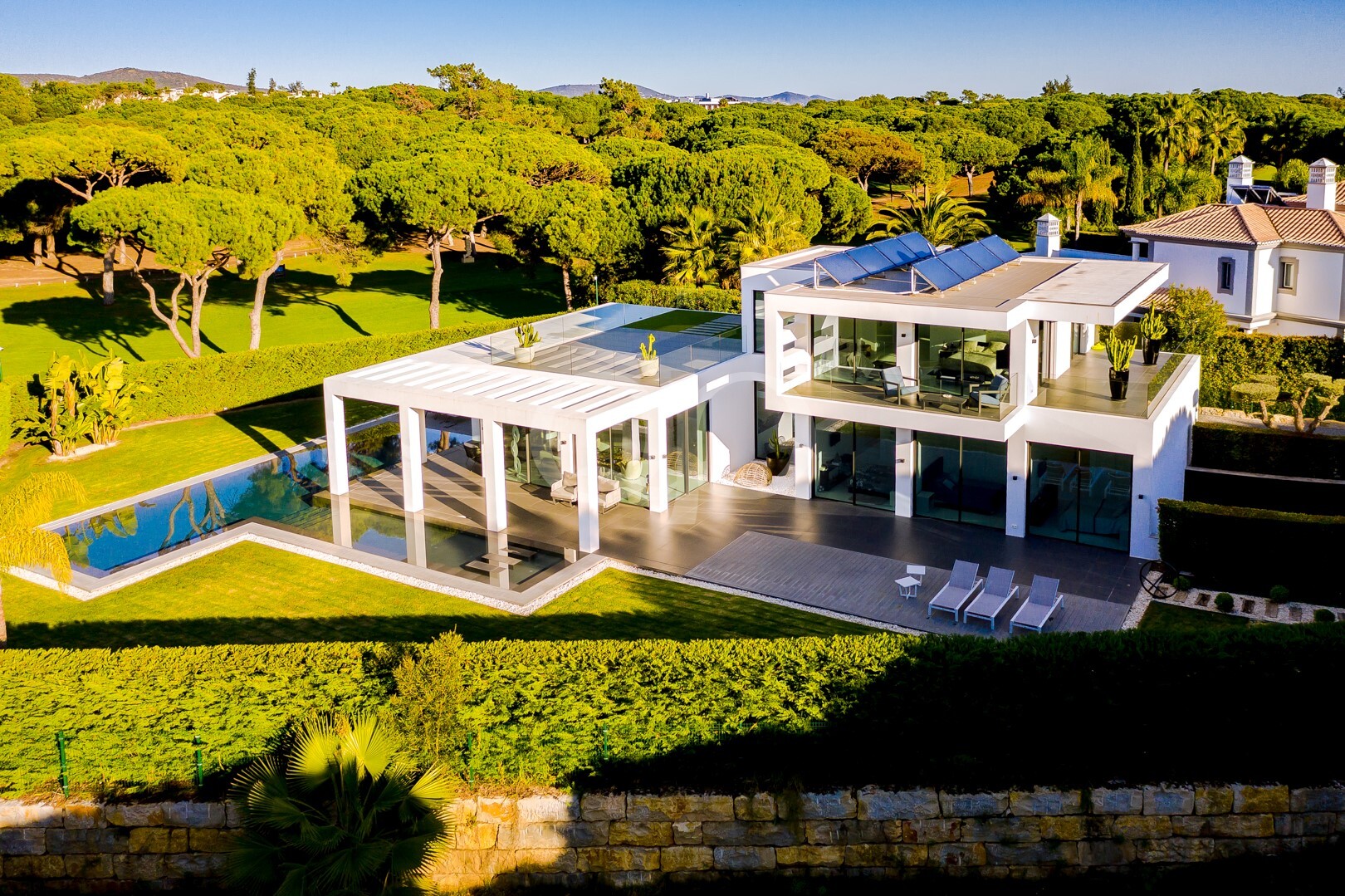 Aerial view of a modern white villa with glass walls, rooftop deck, and long rectangular swimming pool amid manicured grounds and trees.