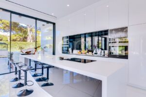 Une cuisine moderne d'un blanc éclatant, dotée d'un long îlot brillant, de tabourets de bar noirs et de grandes portes vitrées donnant sur l'extérieur.