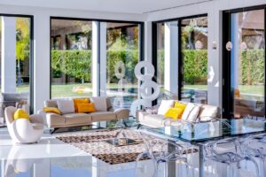 Un salon lumineux et moderne, avec des canapés blancs et des coussins jaunes, une table basse en verre et de grandes fenêtres donnant sur un jardin verdoyant.