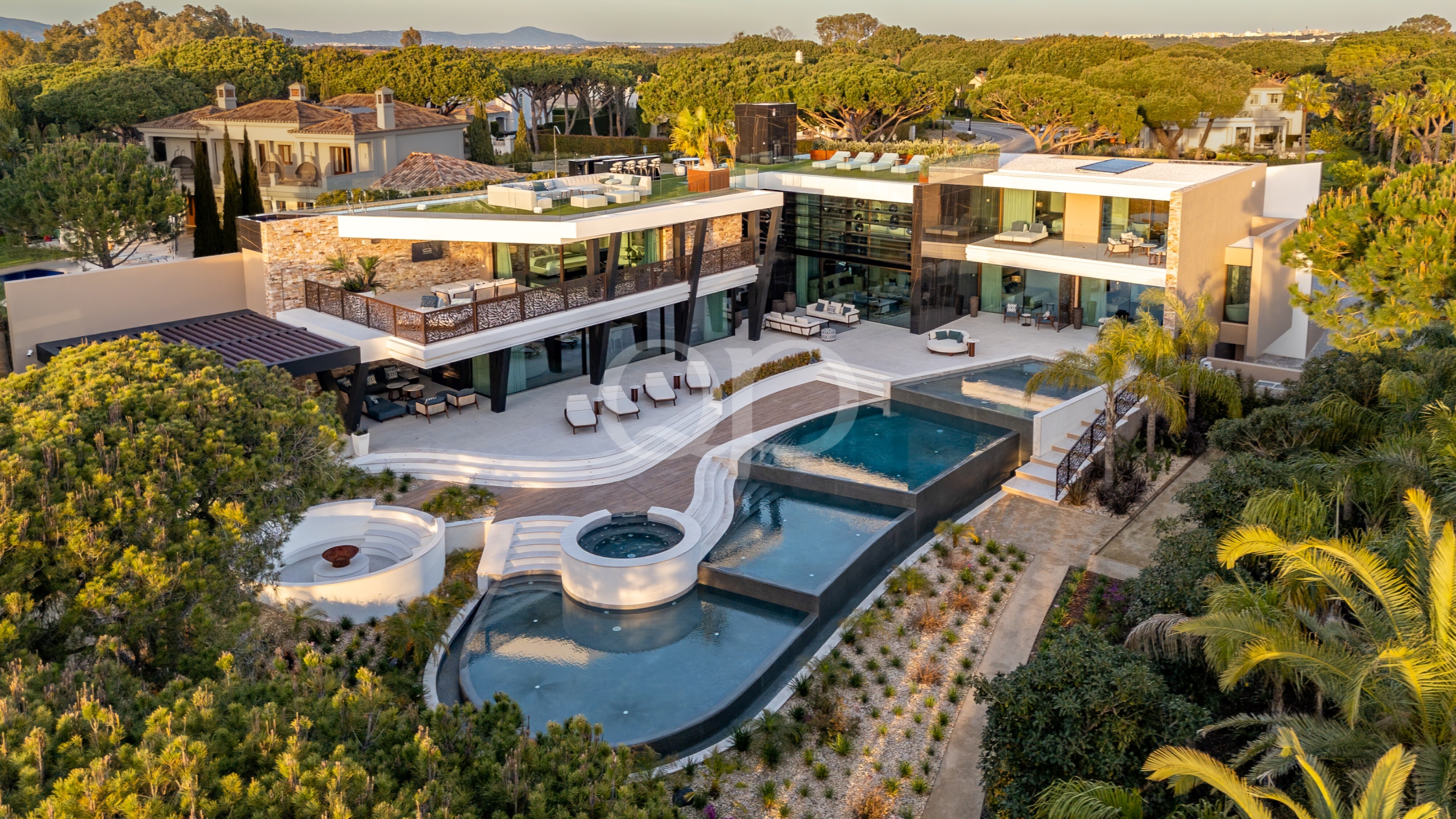 Aerial view of a modern glass-walled villa with multiple terraces, a large swimming pool, spa, and landscaped gardens around the estate.