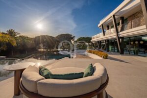 Sunlit modern villa terrace with circular outdoor sofa, decorative metal railing, and a reflecting pool set against palm trees and blue sky.