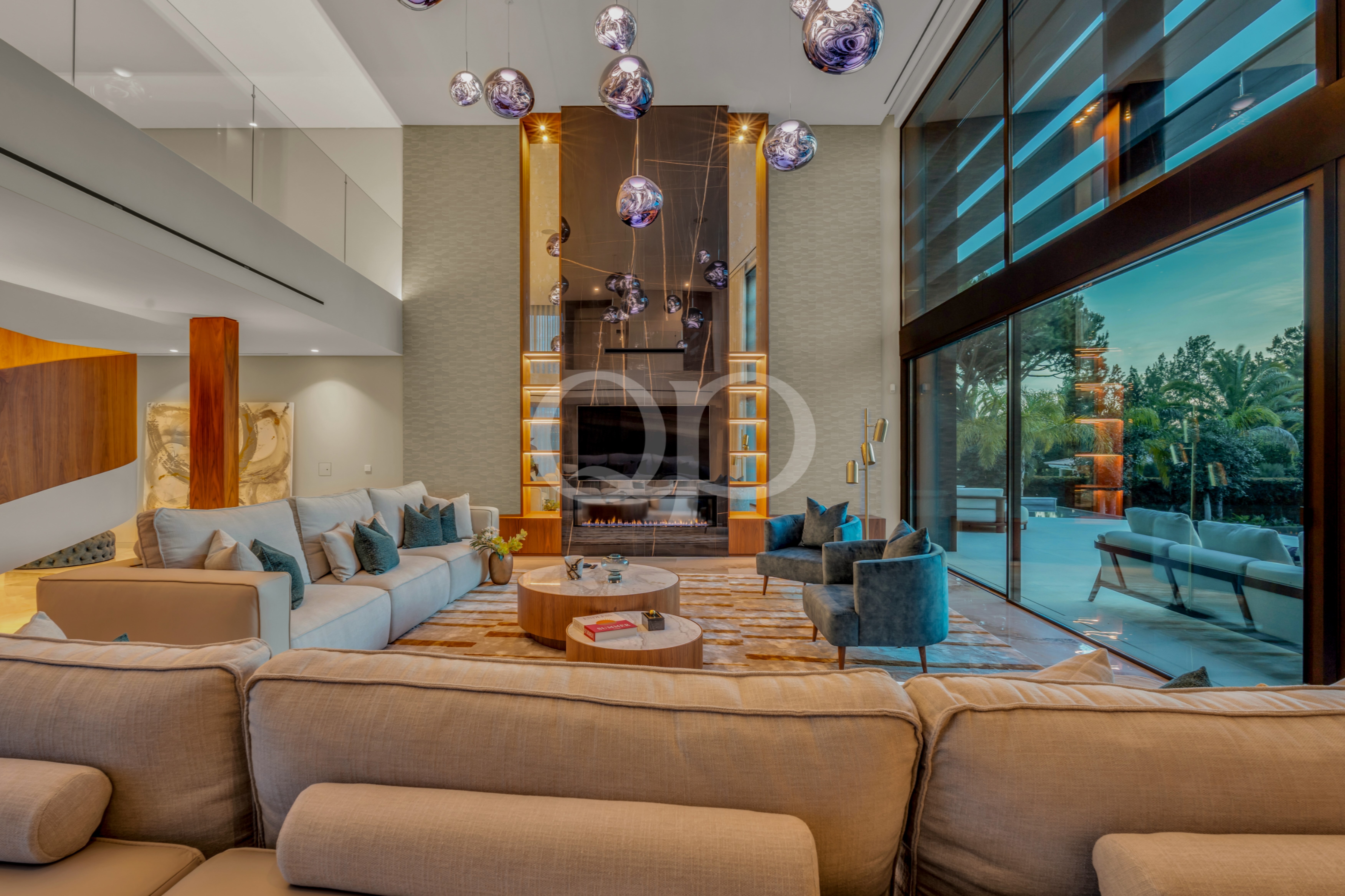 Luxurious living room with a beige sectional, oval marble coffee tables, and a tall fireplace framed by warm wood panels surrounded by glass walls and a view of greenery outside