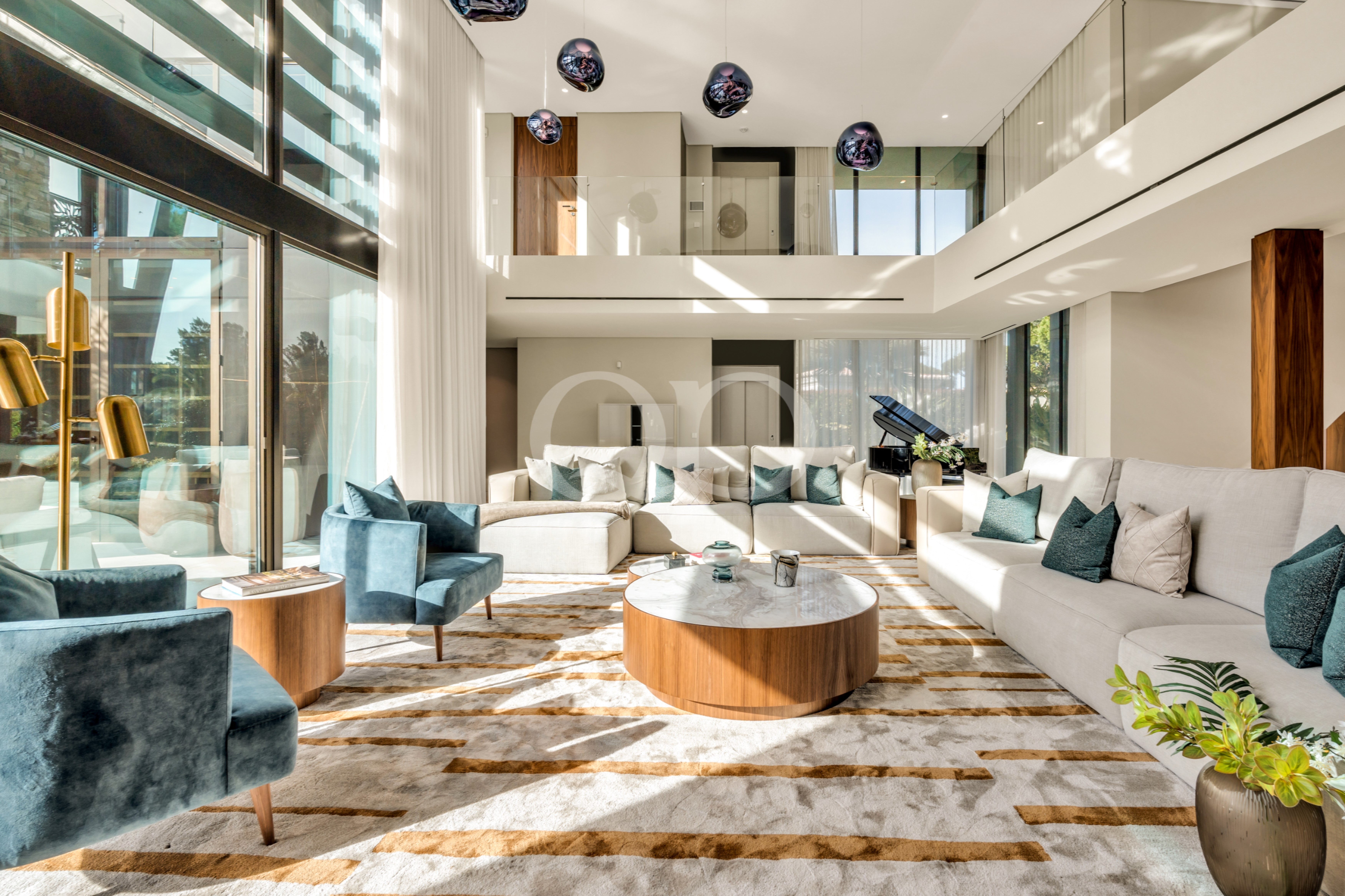 Luxurious open-plan living room with white sectional sofa, teal chairs, marble coffee tables, striped carpet, and large windows letting in natural light.
